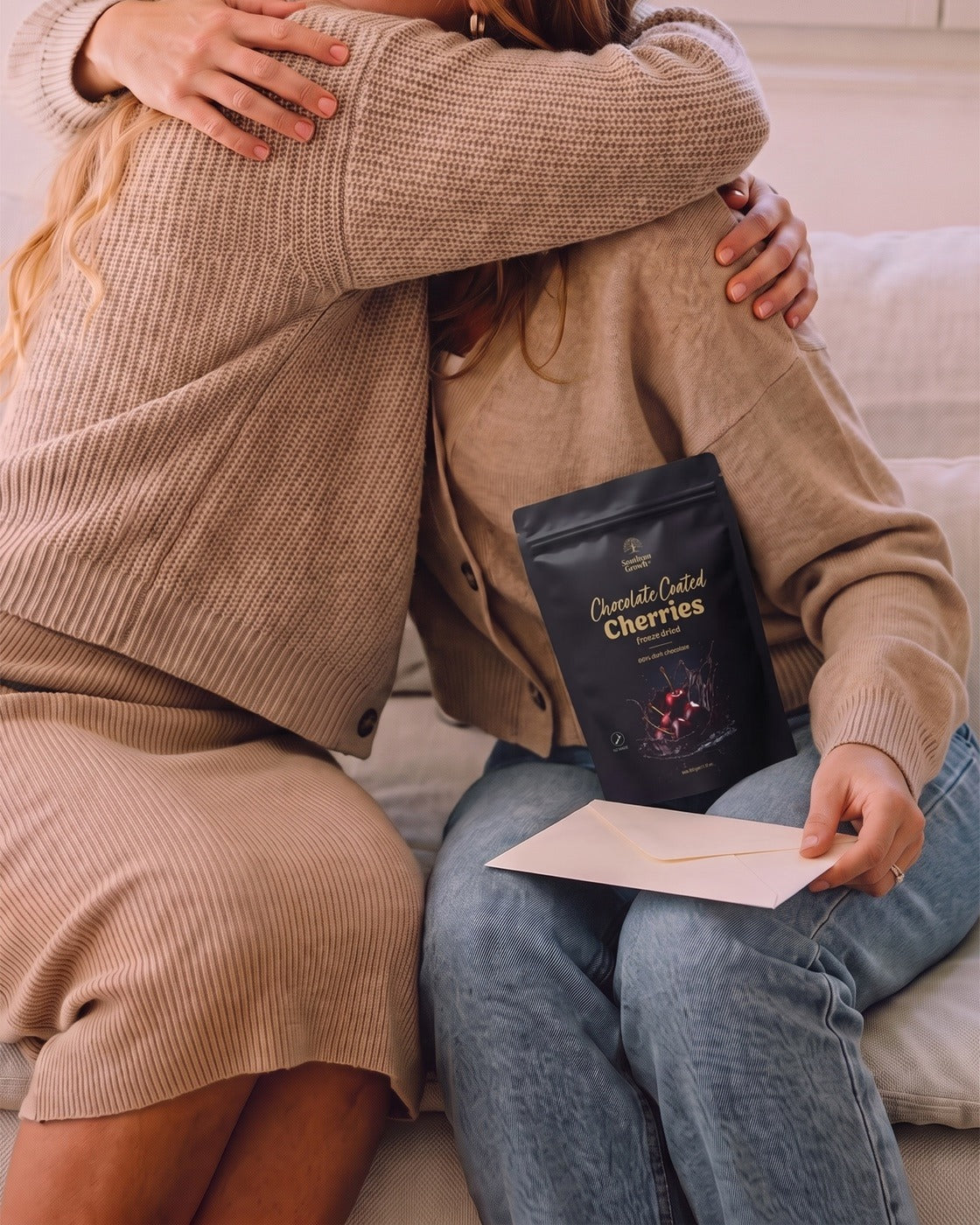 Two people hugging on a sofa, a pack of Chocolate Coated Cherries and a card in hand.