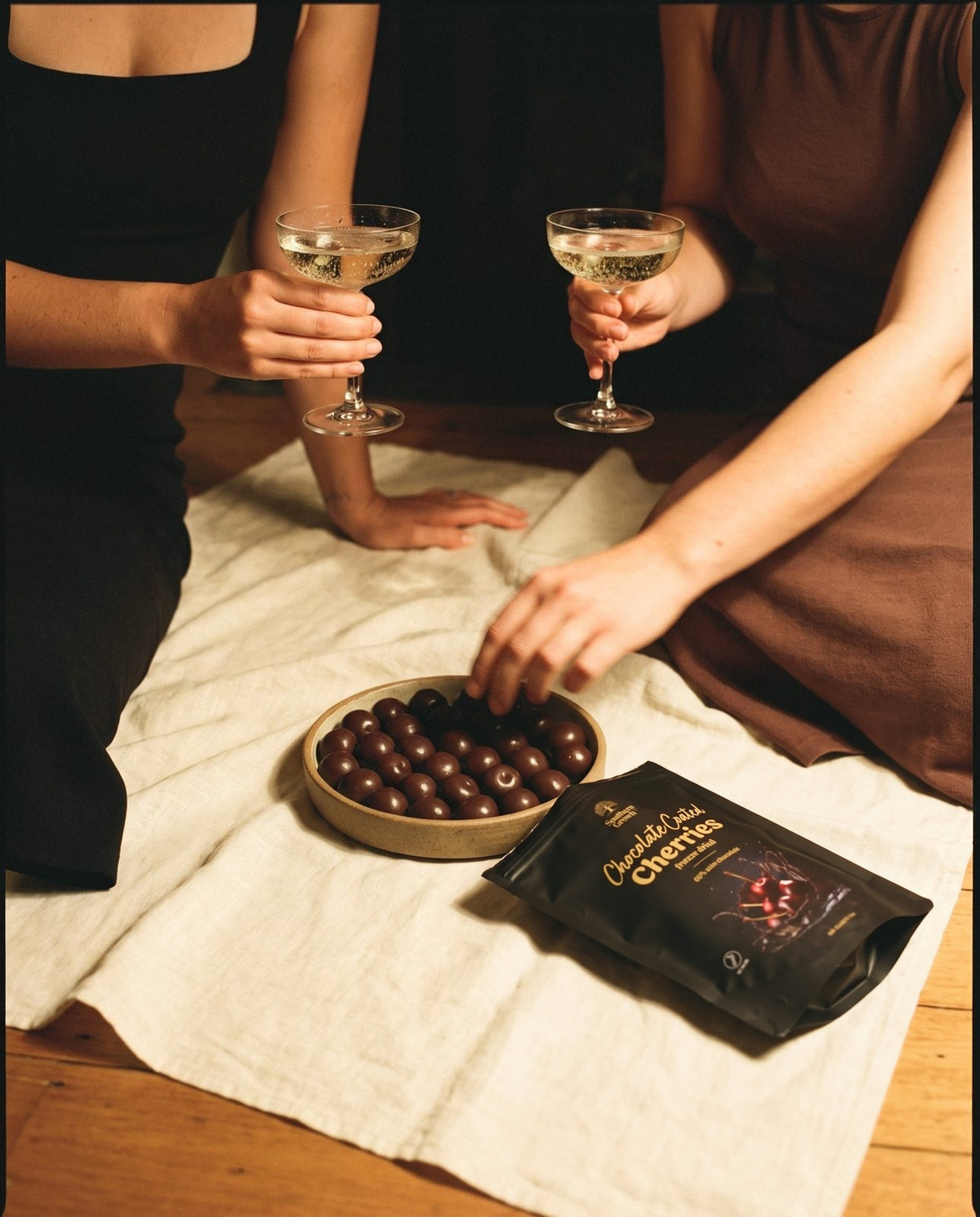 Two friends with glasses of Champagne, sharing a bowl of coated cherries beside a pack.